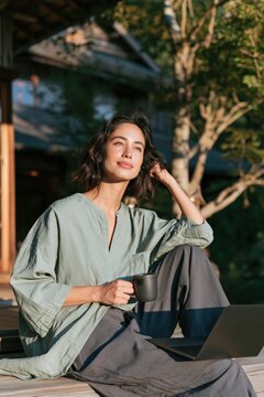 Relaxed woman enjoying morning coffee with laptop on porch in green shirt for remote work lifestyle background and wellness banner design