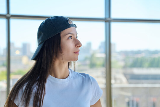 Thoughtful young woman in backward cap looking away by window