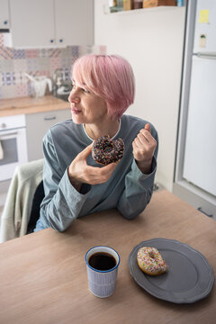 Happy mature woman holds chocolate donut during PMS craving, celebrating dietary relapse and return to sugary comfort food after unsuccessful attempt to resist sweets and control sugar addiction.