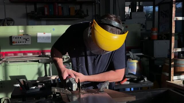 Craftsman wearing a protective face shield carefully grinds a piece of metal with an angle grinder, creating sparks in an industrial workshop