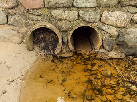 Two rusty pipes emerge from stone wall, releasing murky brown water. Mud and debris surround pipe openings. Scene shows neglected infrastructure near natural setting. Ideal for environmental