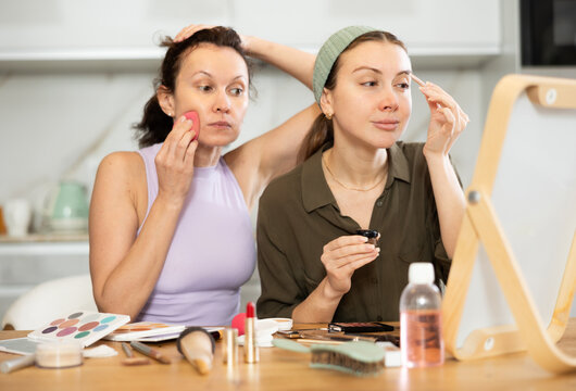 Two focused female friends carefully applying makeup while looking into vanity mirror on table in cozy home setting during daily beauty routine