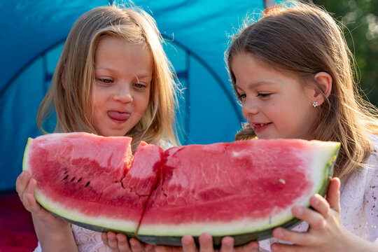 Two cheerful girls holding and eating a large slice of watermelon outdoors during summer camping picnic