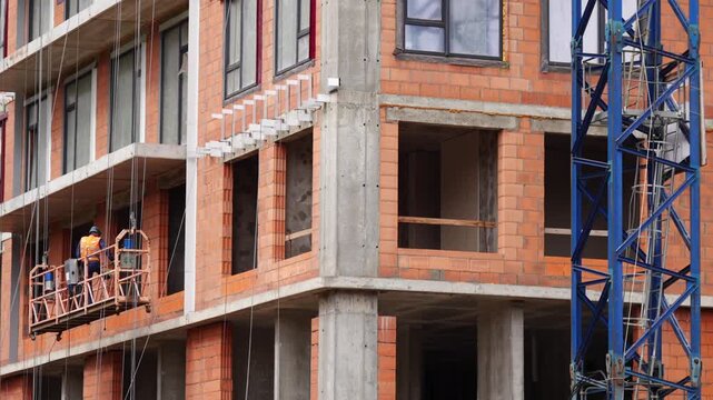 Construction workers on a suspended work platform performing exterior installation on an unfinished multistory building. Active facade construction with exposed concrete structure, masonry walls and t