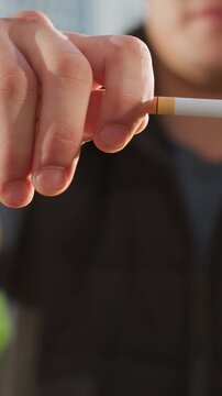 Close up of man holding and lighting cigarette with visible hand details and lit end. background shows urban environment with buildings in soft focus creating depth. Lighting highlights skin