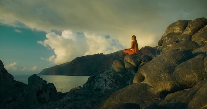 Woman Meditating on Coastal Rock Above Sea at Sunset with Dramatic Sky
