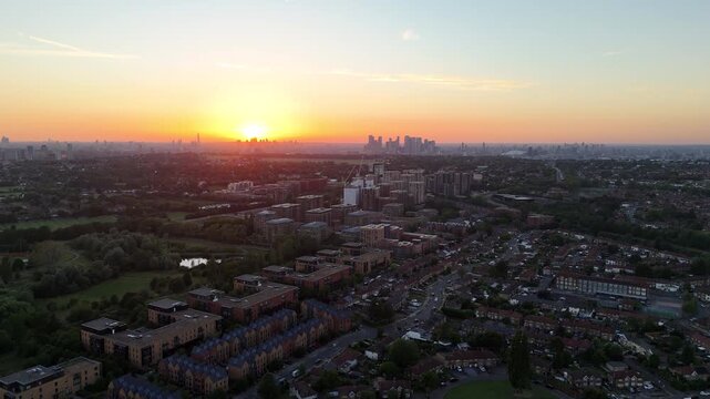Wide aerial view over London at sunset, with residential neighborhoods, green spaces and the distant City of London and Canary Wharf skylines glowing beneath warm evening light.