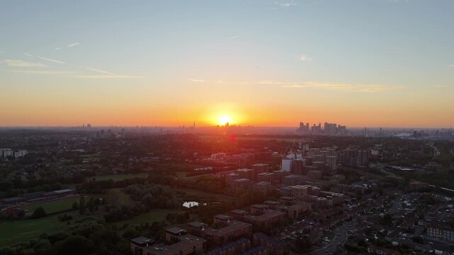 Wide aerial view over London at sunset, with residential neighborhoods, green spaces and the distant City of London and Canary Wharf skylines glowing beneath warm evening light.