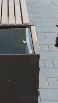 Close up of metal trash can with occasional small pieces of litter inside positioned next to wooden bench on gray brick pavement. Person wearing checkered shirt and jeans briefly appears