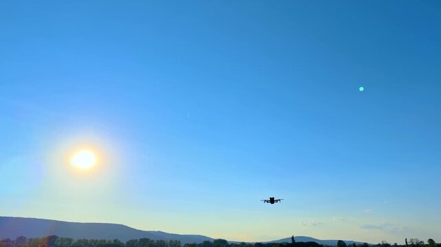 Quadcopter drone flying high in clear blue sky near sun. The small black aircraft is positioned against a bright sun in a gradient sky above far hills.
