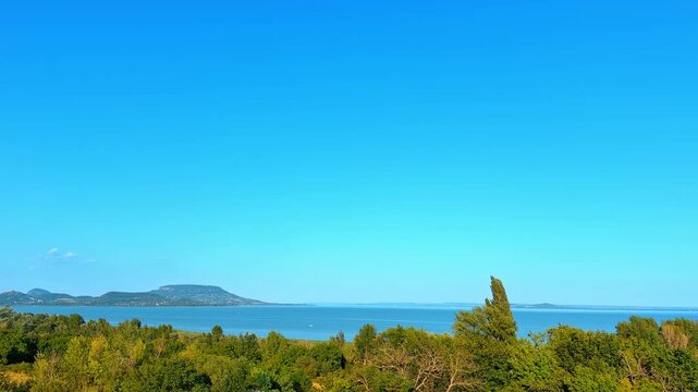 Panoramic view of wide blue lake behind green foliage. Calm water stretches toward distant rolling hills under a clear bright sky during daytime.