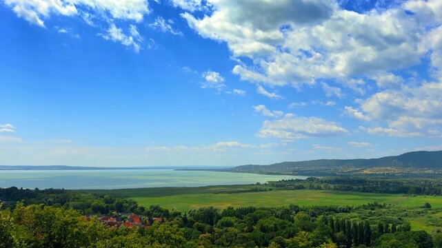 High angle landscape of lake bordered by green fields and hills. Cumulus clouds scatter across a bright blue sky over green vegetation and distant water.