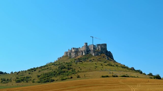 Castle hilltop view, clear sky. A castle sits atop a grassy hill with a crane nearby. The scene shows a bright and clear day.