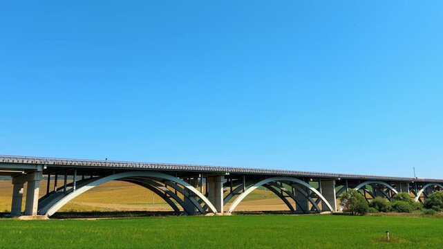 Bridge spans open field under clear sky. A large bridge stretches across a green field on a sunny day. The view shows the bridge's arches and landscape.