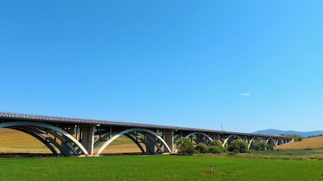 Bridge over green fields, blue sky. A large bridge stretches over green fields. It is a sunny day with few clouds in the sky. The bridge has many arches.