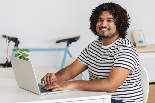 Young adult Indian man smiling while using laptop at desk in home office. Positive remote work environment with natural light. Business concept.