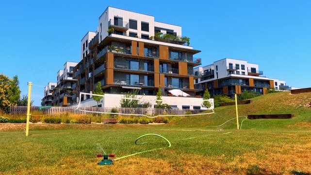 Sprinkler watering grass in urban area. A sprinkler is watering green grass in front of modern buildings during a sunny day. People enjoy their time outside.