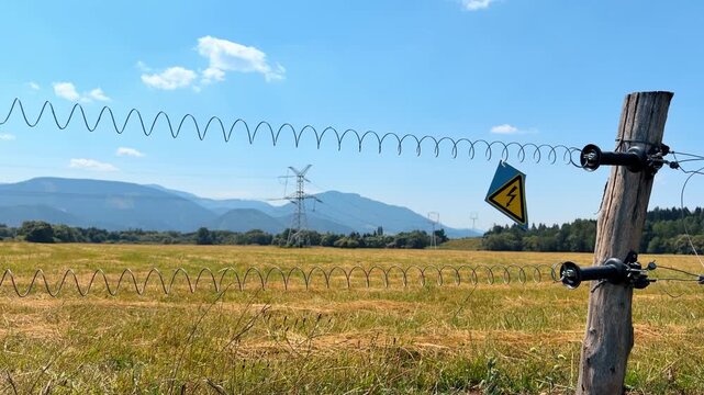 Electric fence by a field and mountains. A field stretches under blue skies near mountains with an electric fence in the foreground and a warning sign.