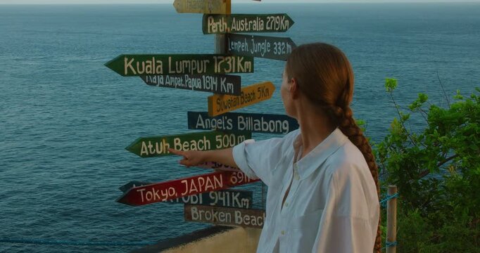 Woman points at colorful wooden signpost showing distances to world cities in Nusa Penida Bali