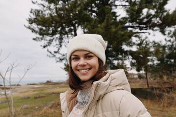 Fototapeta premium Woman in a cozy beige jacket and knit beanie outdoors, smiling with a scenic field and distant water behind her, amid trees and crisp air.