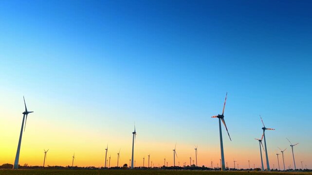 Wind turbines silhouetted against sunset sky. Tall wind generators stand across agricultural horizon under colorful yellow and blue twilight sky.