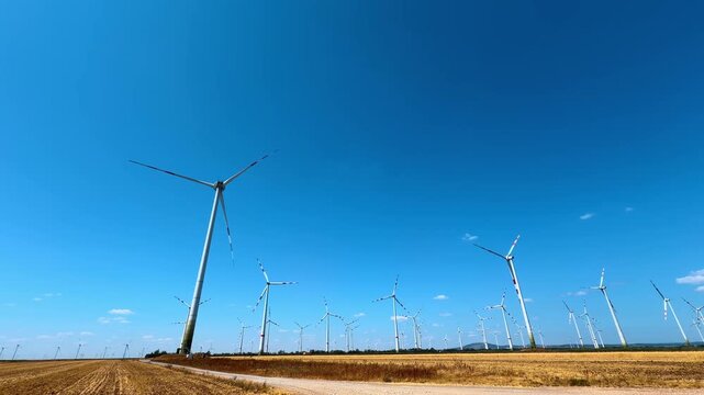 Wind turbines in a blue sky. Wind turbines rotate in a large open field. The sky is bright blue with a few clouds.