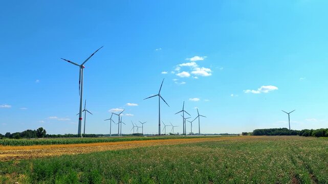 Wind farm across green agricultural fields. Modern wind turbines stand behind lush green field and yellow crops under bright blue sky.