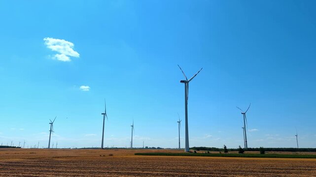 Wind turbines in a field during daytime. Wind turbines stand tall in a wide open field under a clear sky. The sun shines bright as clouds drift by slowly.