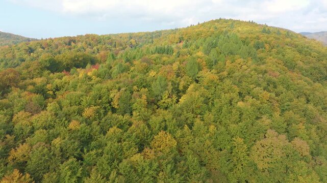 AERIAL: Rolling forested hills, where green, yellow and orange foliage creates a broad autumn mosaic. Layered hills in vibrant colors extend in the distance beneath a bright, lightly clouded sky.