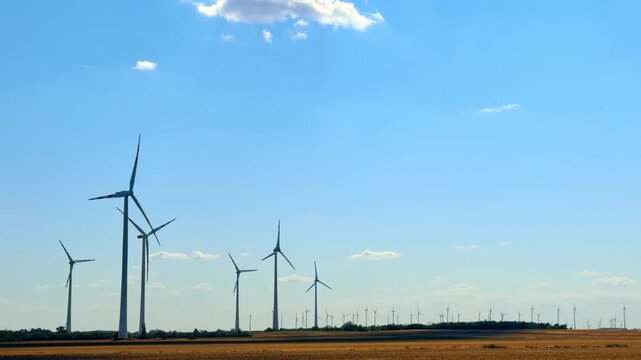 Wind turbines at a farm in the afternoon. Wind turbines spin in a field during the afternoon under a blue sky with clouds. The landscape is open and rural.