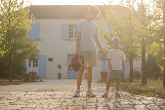 Two boys holding hands and standing on a cobblestone, in outside in front of a house with blue, with cobblestone path, house with blue shutters, potted plants, with white, blue accents.