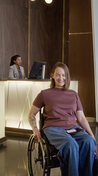 Vertical shot of portrait of Caucasian young adult woman with disability in wheelchair holding passport and smiling in hotel lobby. Clip promoting inclusive travel and accessible tourism