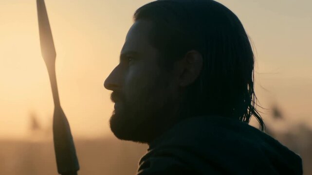 Bearded warrior holding spear in warm sunset light with soldiers blurred behind