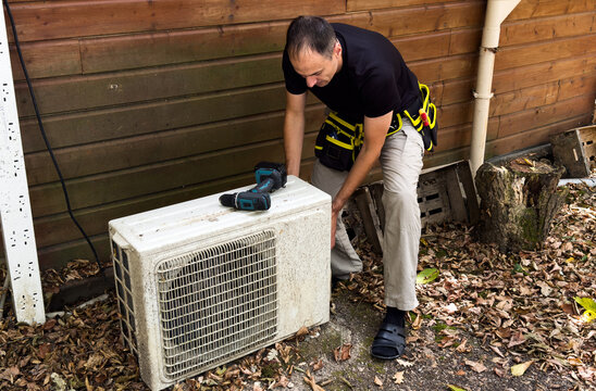 HVAC technician services an outdoor unit near a house wall, holding a tool. Concept of home heating, cooling and maintenance.