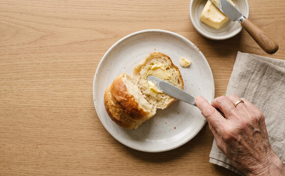 Elderly woman spreading butter on fresh bread roll at wooden table top view
