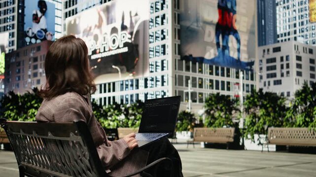 Programmer writes code on a laptop while sitting on a park bench outdoors; the developer uses a programming software interface on the notebook screen in a city environment while working remotely.