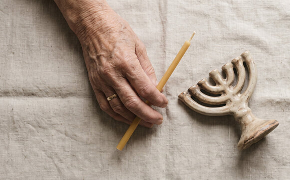Elderly female hand holding a yellow candle next to a ceramic menorah on linen fabric