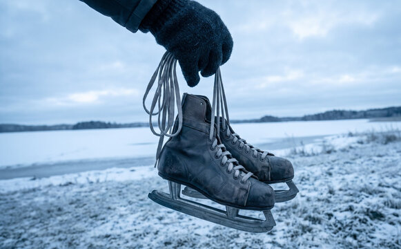 Person holding vintage leather ice skates by the laces near a frozen lake in winter landscape