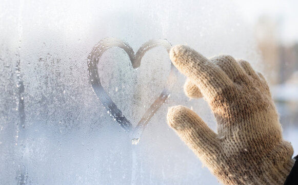 Person in warm mitten drawing a heart shape on a frosted window during winter season representing love