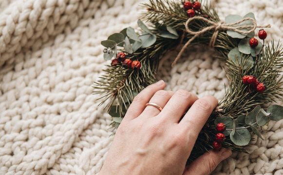 Woman touching handmade Christmas wreath with pine branches and red berries on cozy knitted blanket