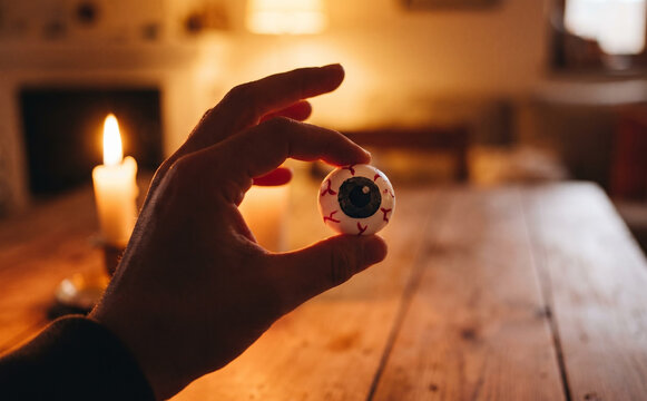 Spooky Halloween concept featuring a hand holding a fake eyeball over a rustic wooden table lit by a candle in a dark room