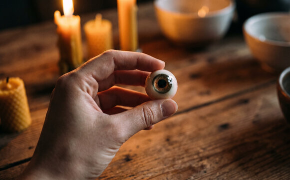 A hand holding a realistic prosthetic eyeball on a rustic wooden table with burning candles for a spooky Halloween concept