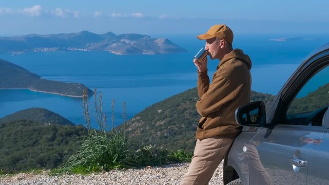 Man drinking coffee and enjoying the scenic mountain sea view. Relaxed man leaning against his car, drinking a hot beverage from a mug while enjoying the beautiful ocean during road trip stop