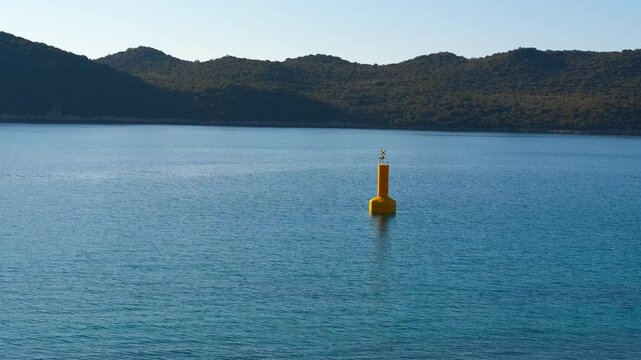 Yellow navigational buoy floating in a tranquil sea bay. Calm blue sea with a single yellow navigational buoy gently bobbing on the water surface and a tranquil, green coastline in the background