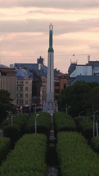 An aerial view showcases the Freedom Monument rising above a tree lined boulevard in Riga, Latvia during a peaceful sunset.
