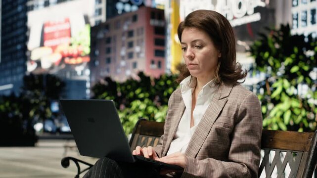 Businesswoman types on laptop keyboard and drinks coffee on park bench
