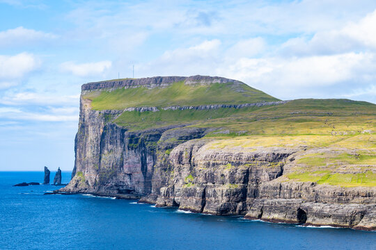 Distant telephoto view of towering sea cliffs and grassy plateaus along the coastline, with the sea stacks Risin og Kellingin visible offshore, Eidiskollur, Eysturoy, Faroe Islands, Denmark