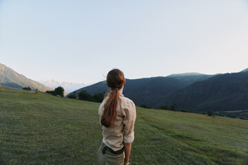 Young woman with long hair stands on green hilltop looking at mountain range under clear sky, enjoying peaceful nature and outdoor lifestyle moment.
