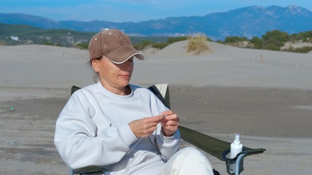 Woman knitting outdoors while enjoying a sunny day. Woman wearing a baseball cap sitting on a camping chair outdoors, enjoying a relaxing day while knitting with yarn and needles against a backdrop
