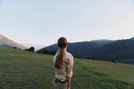 Young woman with long hair stands on green hilltop looking at mountain range under clear sky, enjoying peaceful nature and outdoor lifestyle moment.